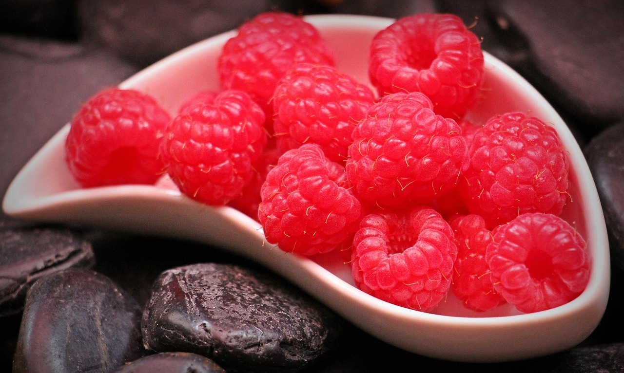 A pale pink ceramic bowl filled with red raspberries beside dark stones on a deep brown surface.