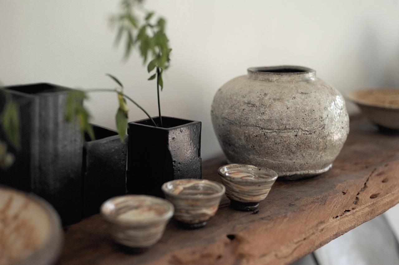 A rounded gray ceramic vase and several small cups sit on a rustic wooden shelf beside dark planters and a thin plant.