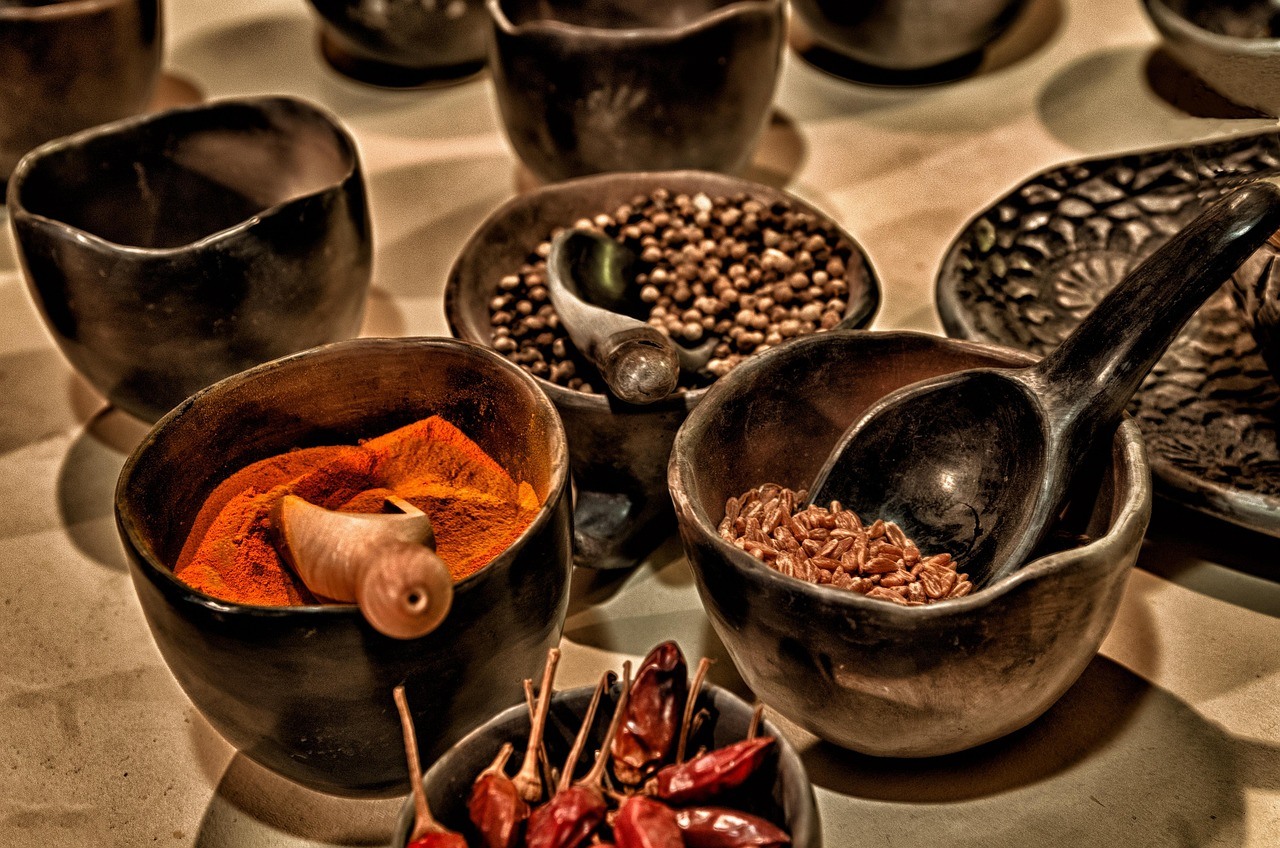 Dark ceramic bowls and cups hold red powder, seeds, dried chilies, and utensils on a warm patterned table.