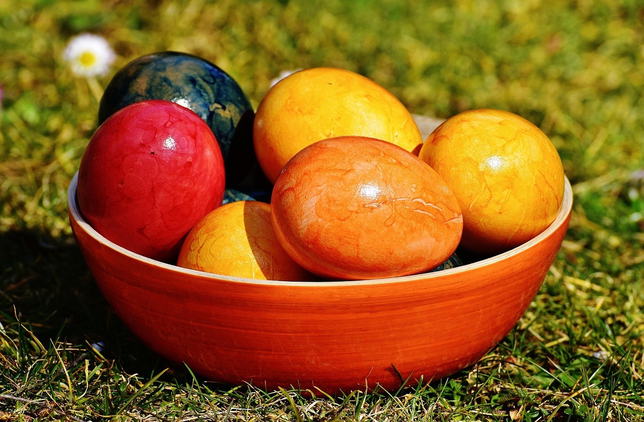 A wide orange ceramic bowl holds glossy colored spheres on sunlit grass.